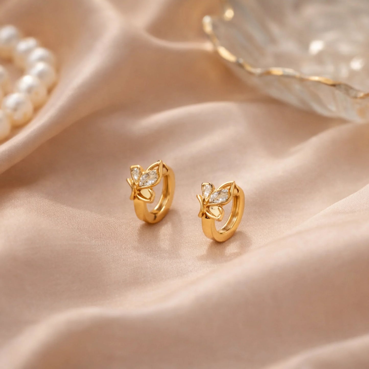 Butterfly gold huggie earrings styled on satin fabric with pearl accents
