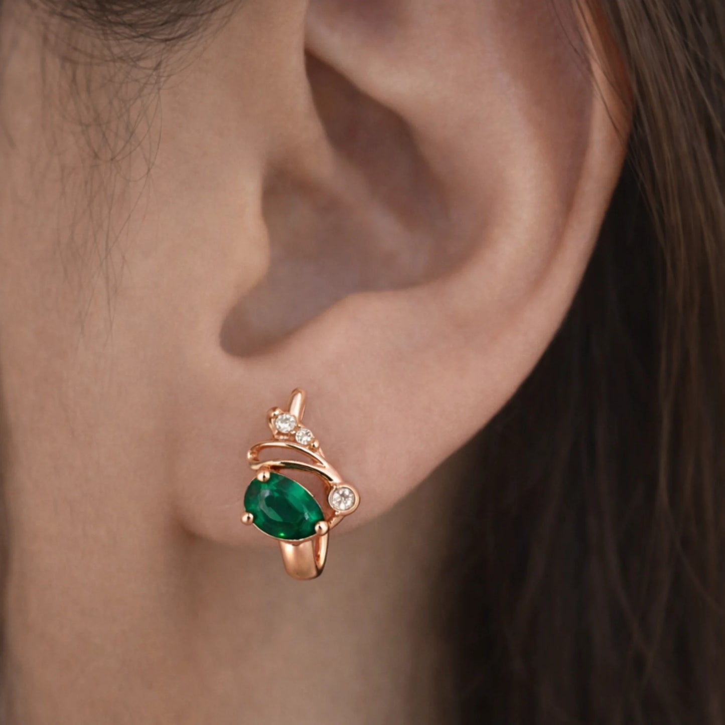 Rose gold huggie hoop earrings with green stone and diamonds worn on ear close-up view