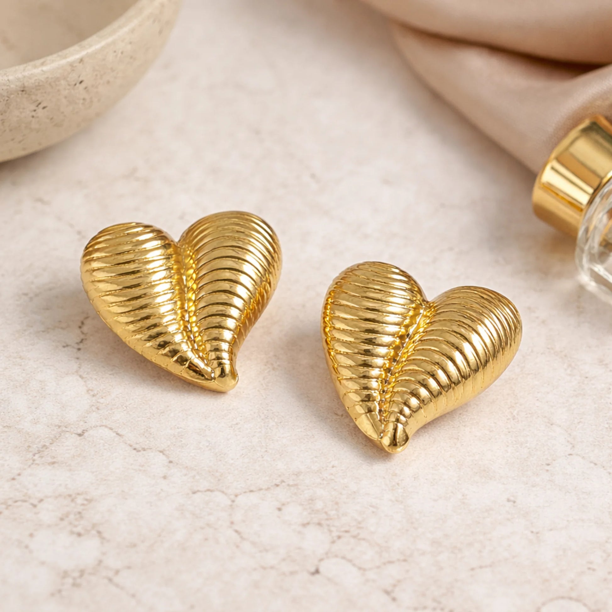 Gold heart-shaped earrings on a light surface with a bottle of perfume.