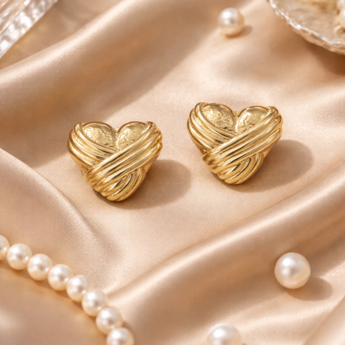 Gold heart-shaped earrings on a soft pink fabric background with pearls.