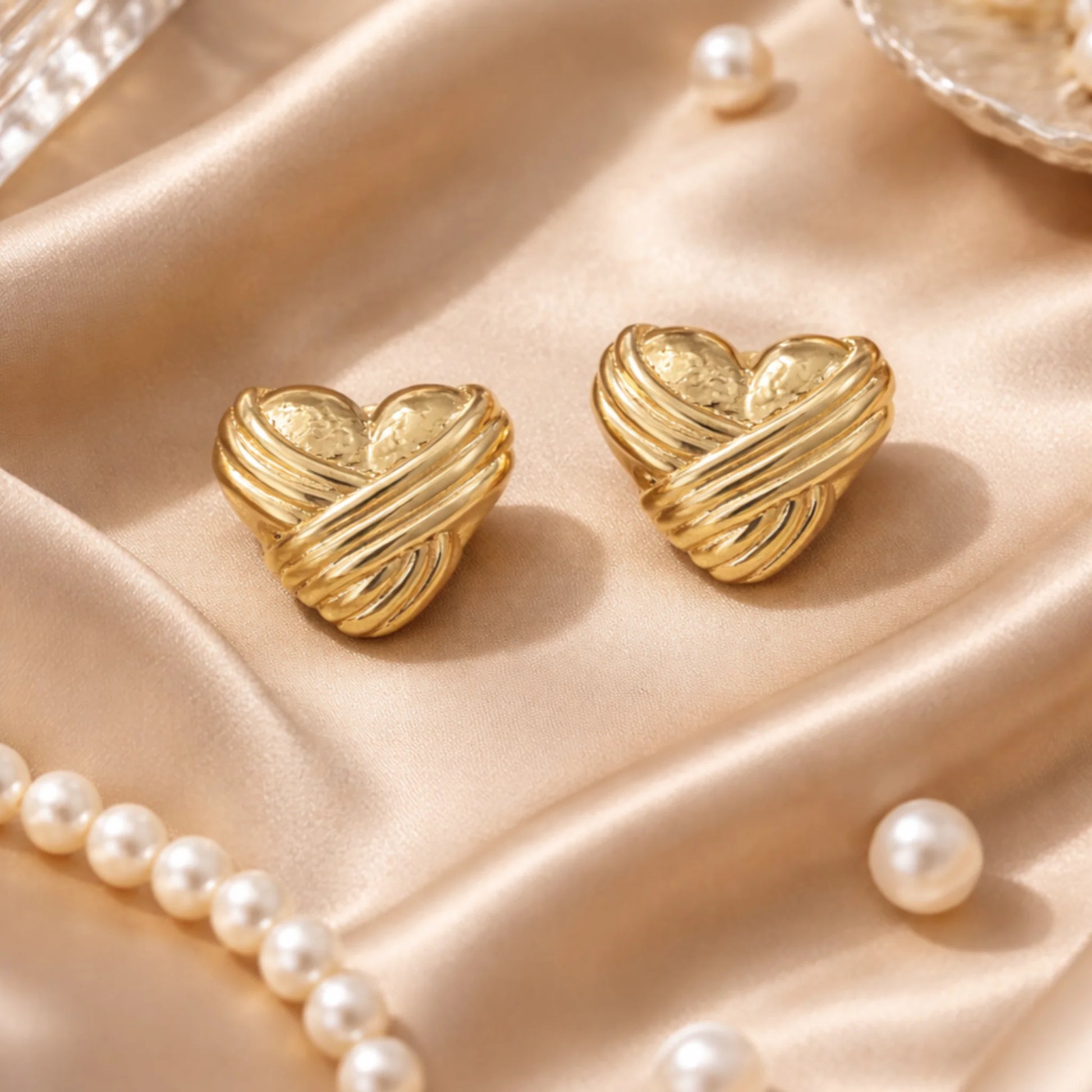 Gold heart-shaped earrings on a soft pink fabric background with pearls.