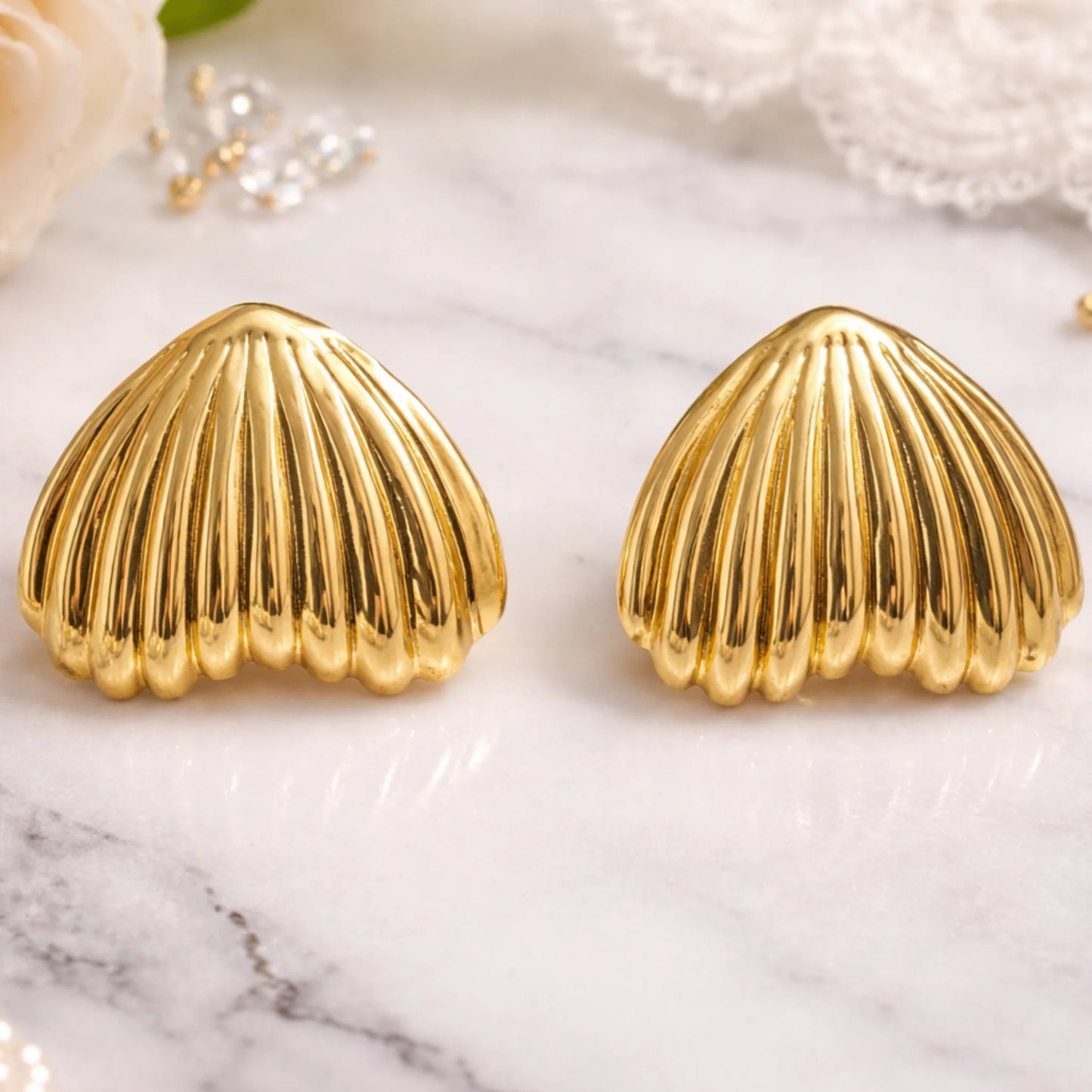Gold shell-shaped earrings on a marble surface with pearls and flowers.