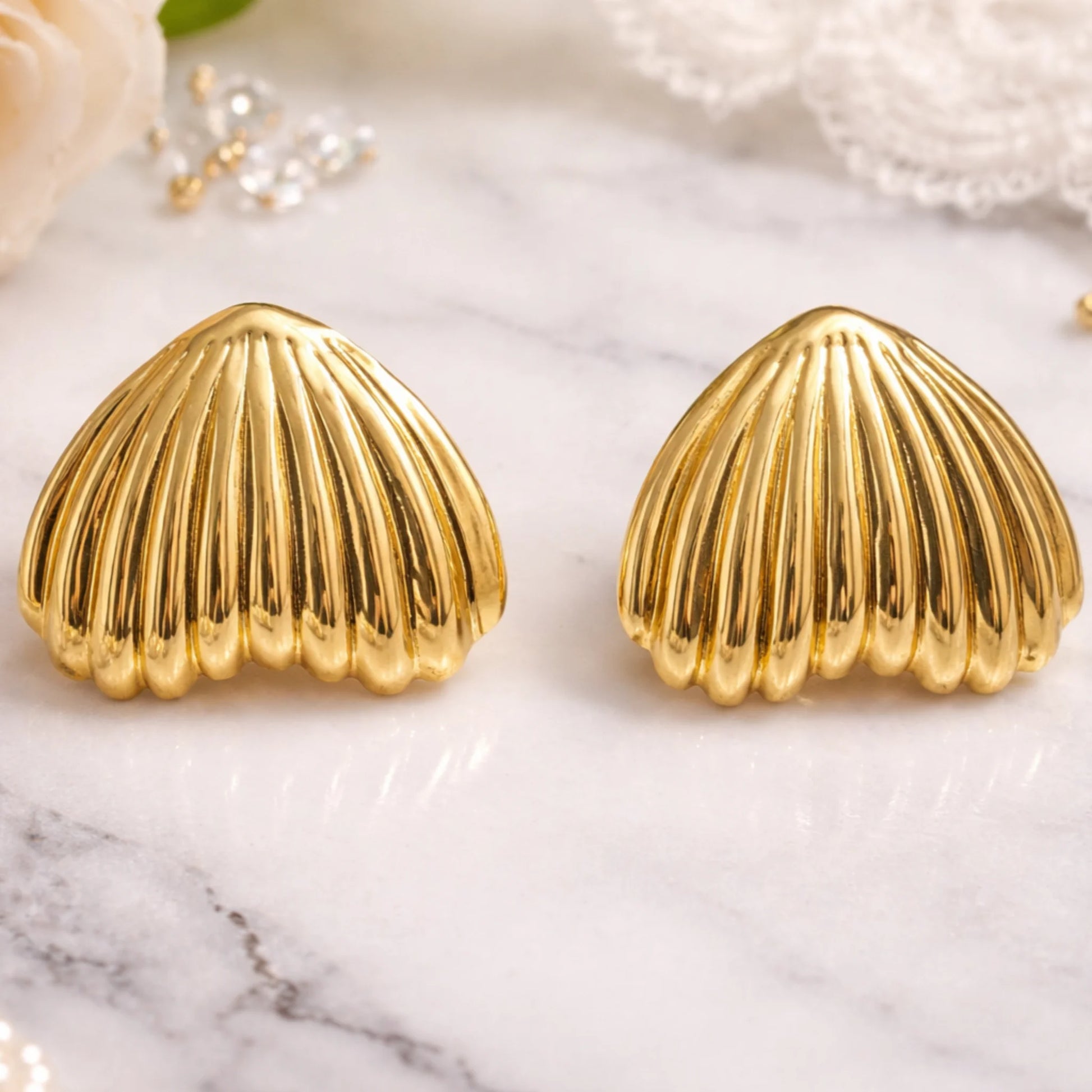 Gold shell-shaped earrings on a marble surface with pearls and flowers.