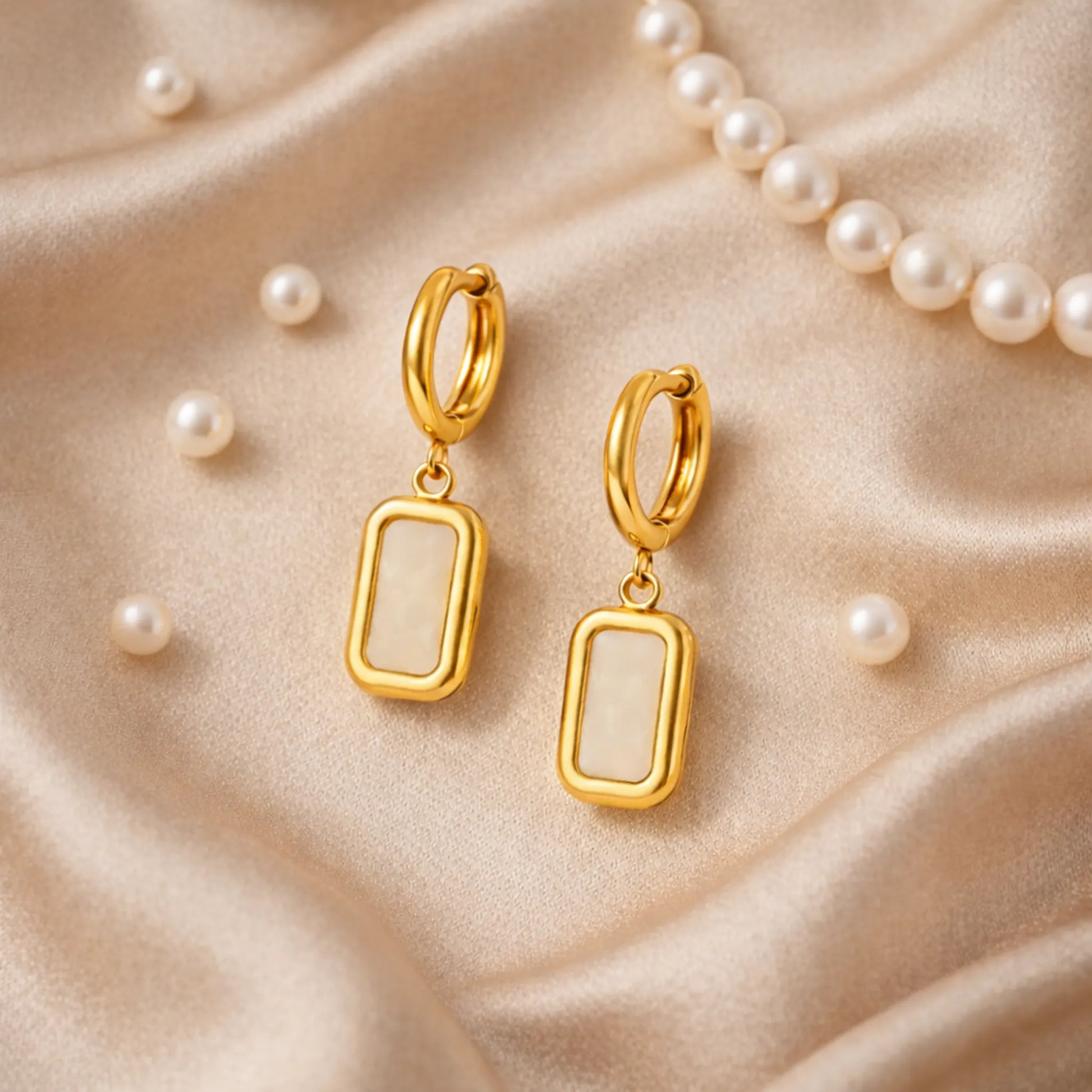 Gold hoop earrings with rectangular drops on a beige fabric background with pearls.