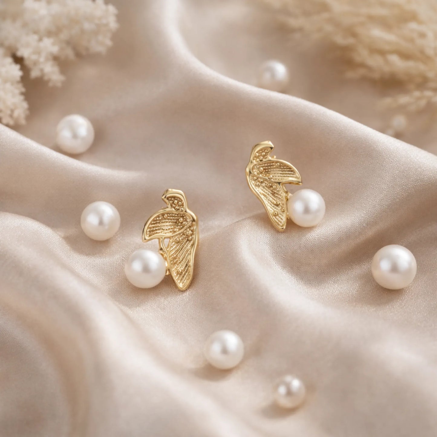 Gold plated pearl leaf earrings by YUBIRAA displayed on soft satin fabric with scattered pearls, showcasing elegant anti tarnish gold polish finish