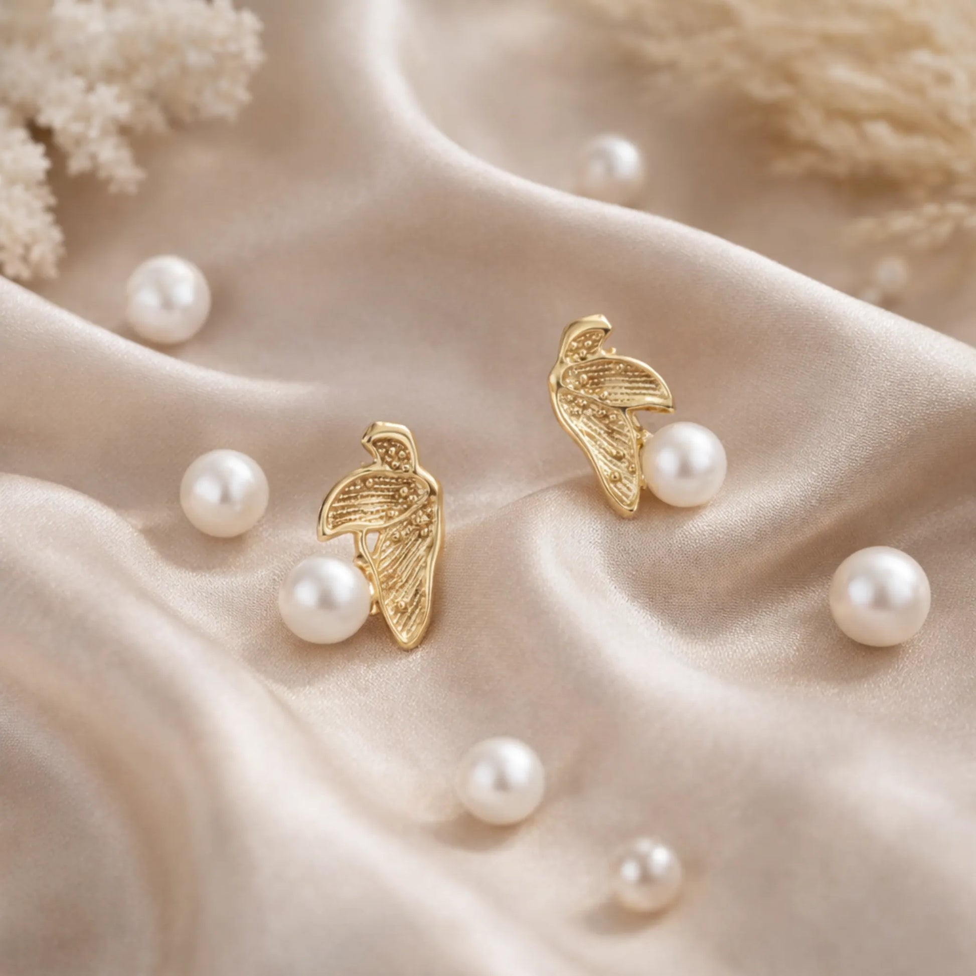 Gold plated pearl leaf earrings by YUBIRAA displayed on soft satin fabric with scattered pearls, showcasing elegant anti tarnish gold polish finish