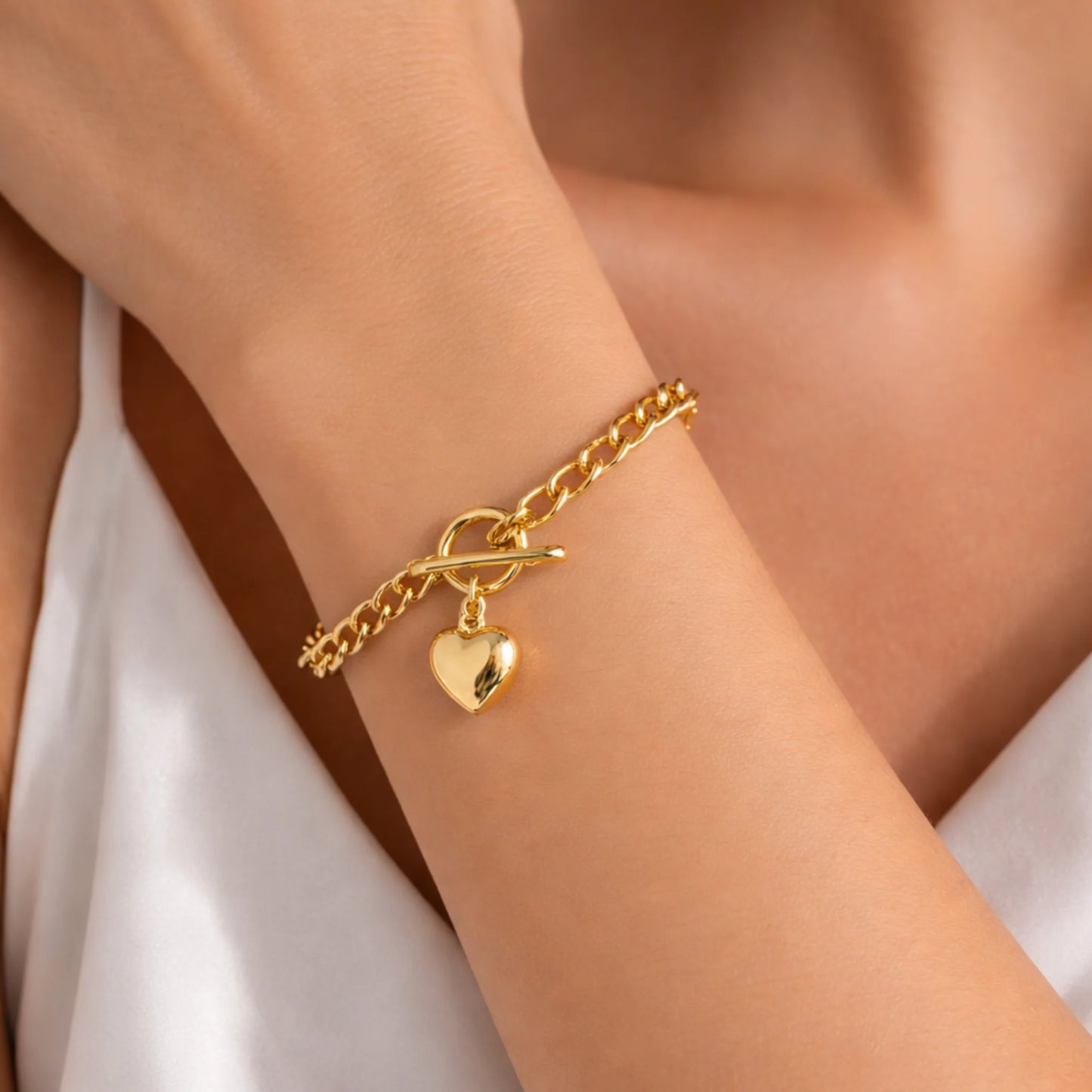 Gold chain bracelet with a heart charm on a wrist against a neutral background