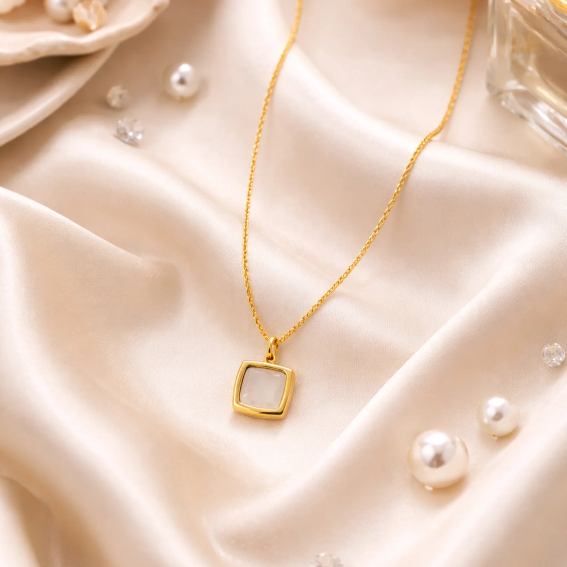 Gold necklace with a square pendant on a beige satin surface with pearls.