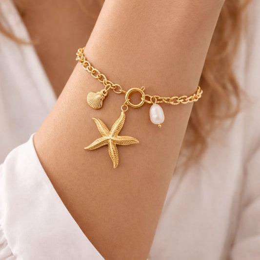 Gold bracelet with starfish, shell, and pearl charm on a wrist.